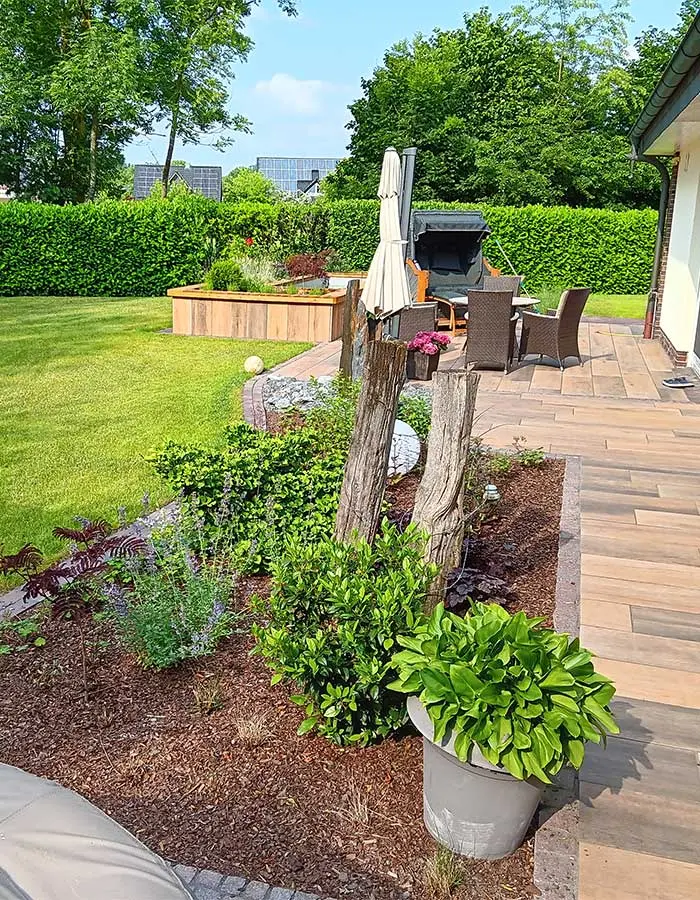 Garten mit Holzstumpf, verschiedenen Pflanzen und einer Terrasse mit Möbeln.